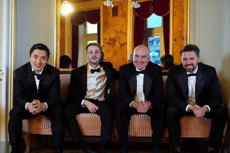 Die vier Sänger lächelnd in Reihe vor goldenem Spiegel